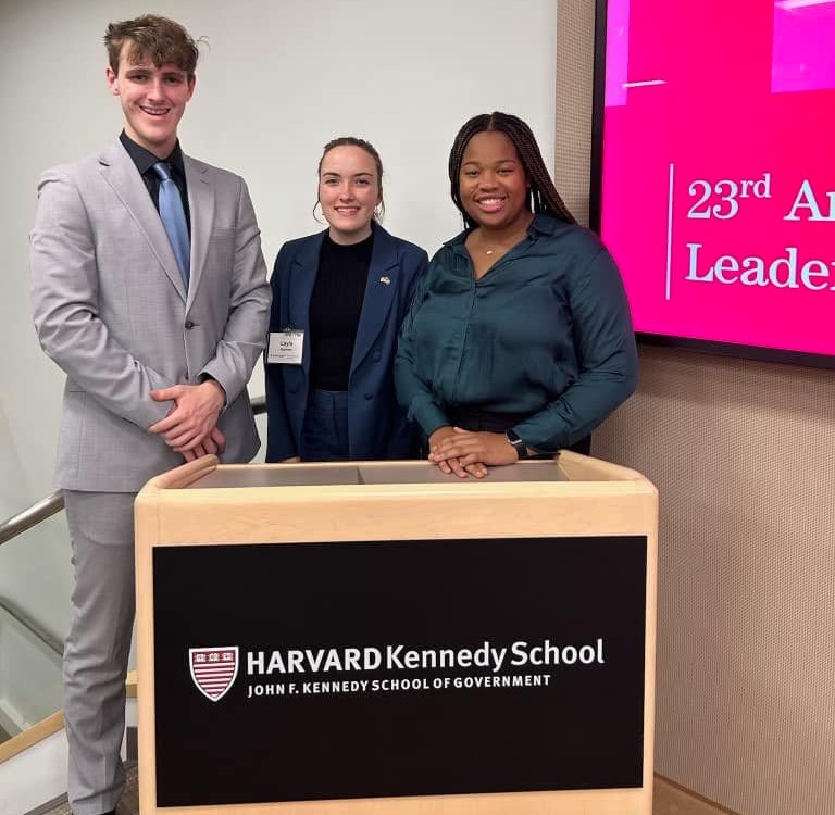 Three students stand at a podium that has a Harvard sign on it. 